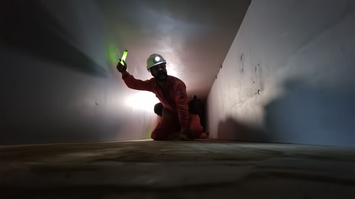 A man in orange construction worker garb and a helmet with a lamp on it holds up a green glowstick as he crawls through a white concrete tunnel in a very grainy scene from the V/H/S/85 segment “God of Death.”