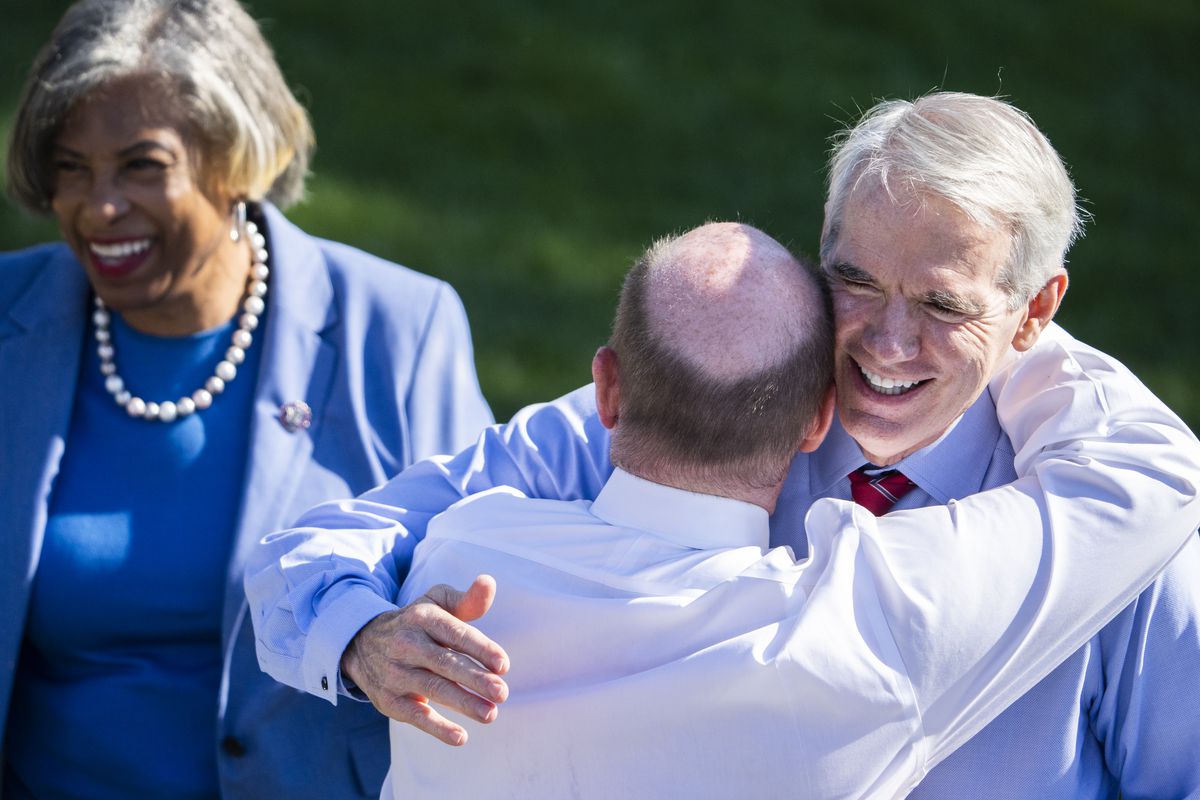 Coons, seen from behind, throws an arms around Portman’s neck. Both have doffed their blazers and are in shirt and tie. Portman, his white hair blowing in the wind, smiles broadly.