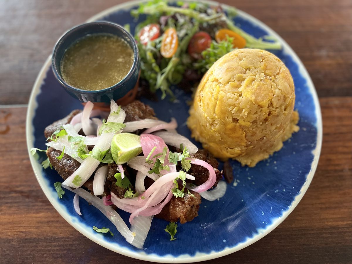 A mound of bright mofongo beside pork buried beneath pickled onions, a salad, and side of green sauce.