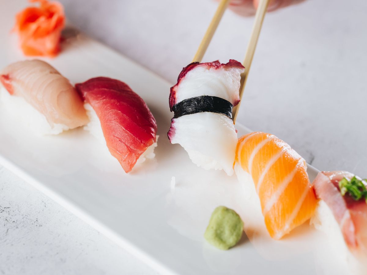 Five pieces of nigiri are on a long white plate. A hand holding chopsticks picks one of them up. At Zen Sushi.