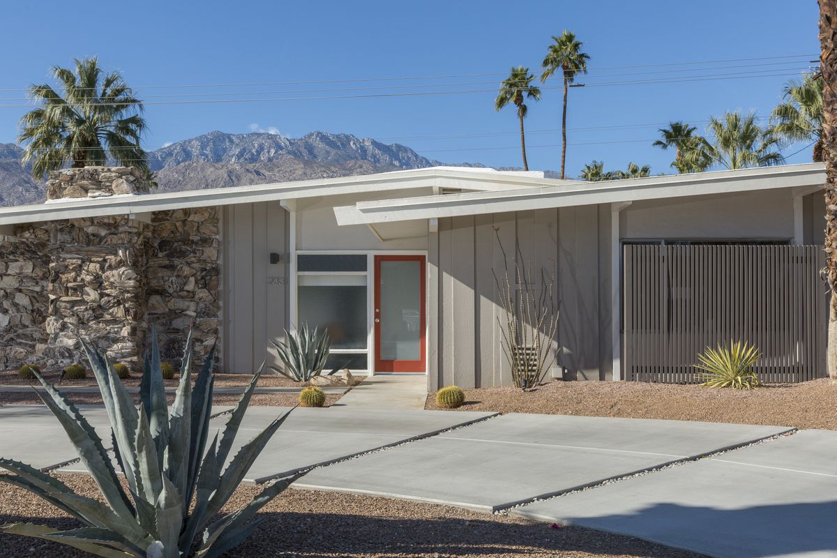Exterior shot of front of single-story house with slanted roof, brick detailing and grey, wood-paneled walls with red trimmed front door. Desert landscaping. 