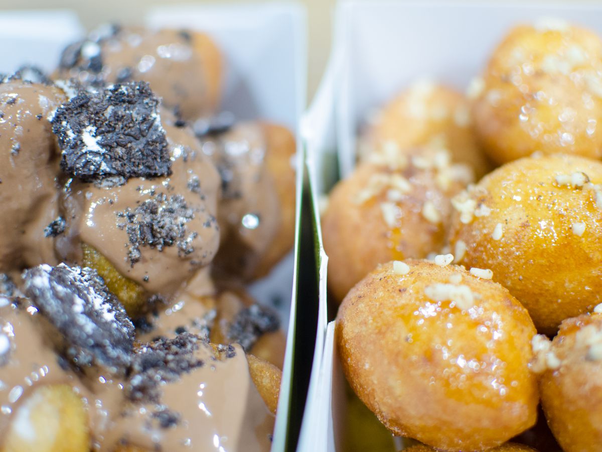 Closeup shot of two side-by-side paper containers of fried dough balls. One is topped in a chocolate glaze with crushed Oreos.