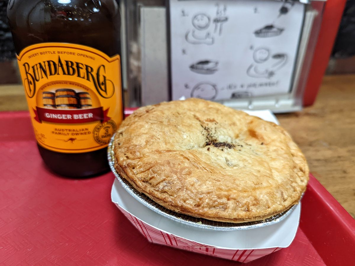A small round savory pie sits on a red tray with a brown glass bottle of Bundaberg ginger beer visibile in the background.