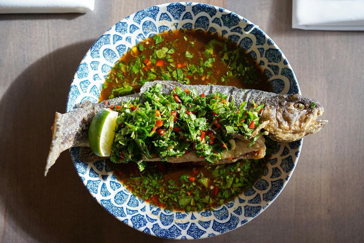 A whole fried trout covered in chopped cilantro and chili peppers over a bowl of brown sauce.