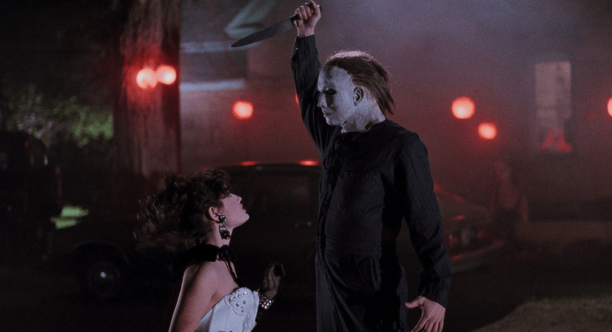 A shaggy Michael Myers stiffly extends his arm above his head, ready to stab a kneeling woman in a white dress, in Halloween 5: The Revenge of Michael Myers.