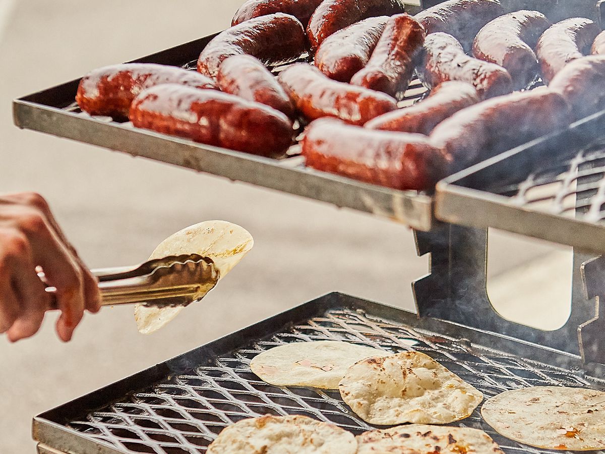 A cook moves a tortilla with tongs on one level of a grill, while sausages roast on another level.