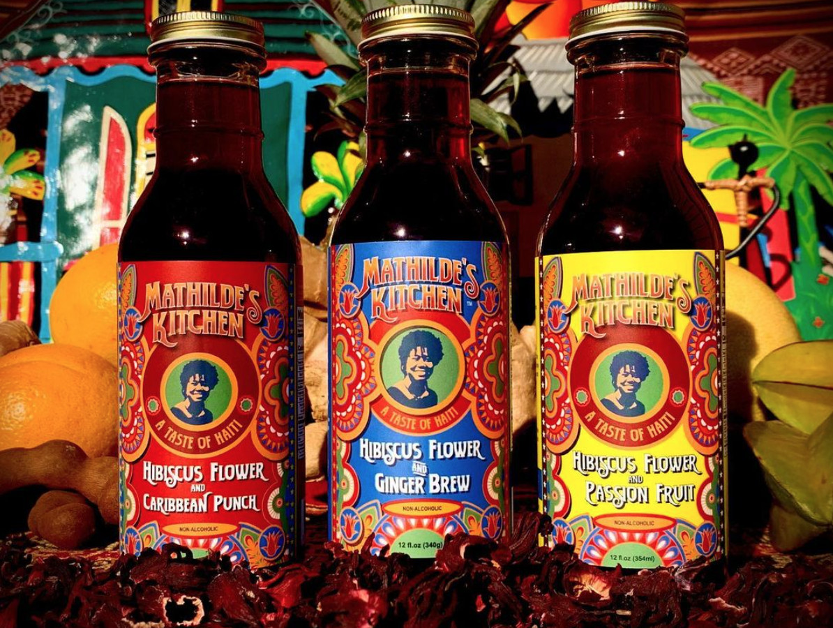 Three colorful bottles of beverages from Mathilde’s Kitchen sit in a row, in front of bananas and oranges. The flavors are (left to right) Hibiscus Flower and Caribbean Punch, Hibiscus Flower and Ginger Brew, and Hibiscus Flower and Passion Fruit.