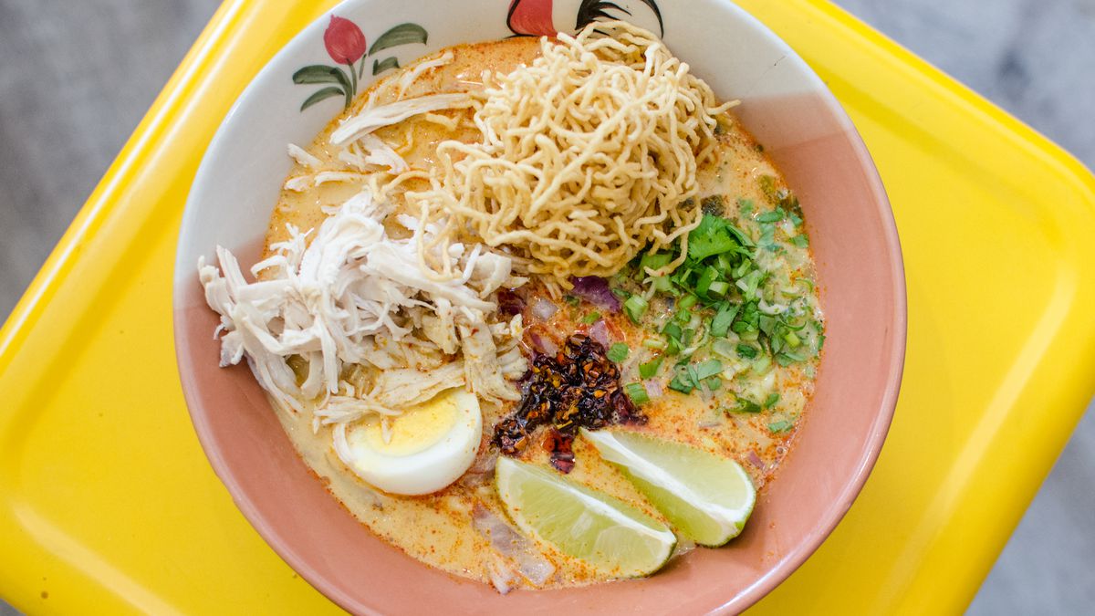Overhead shot of khao soi on a bright yellow surface. The bowl has a traditional Thai pattern on it, including a rooster. The khao soi includes pickled mustard greens, an egg, lime wedges, a nest of crispy noodles, and more.