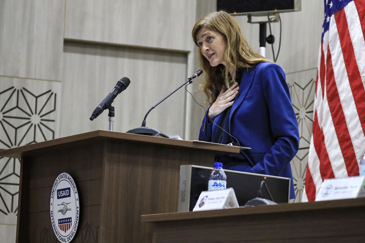 USAID Administrator Samantha Power, wearing blue and with a US flag behind her, gestures as she speaks into a microphone on a brown wood podium.