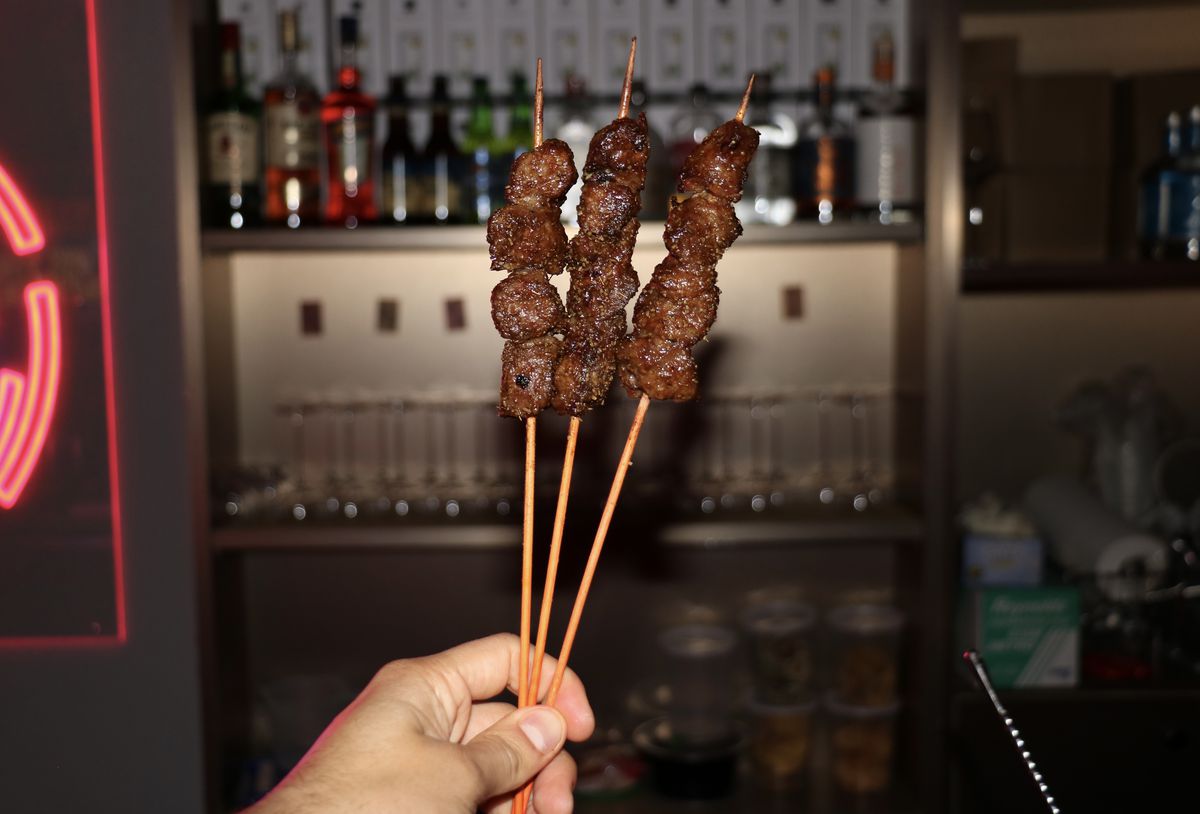 A hand holds three short rib skewers up against a dimly lit bar.