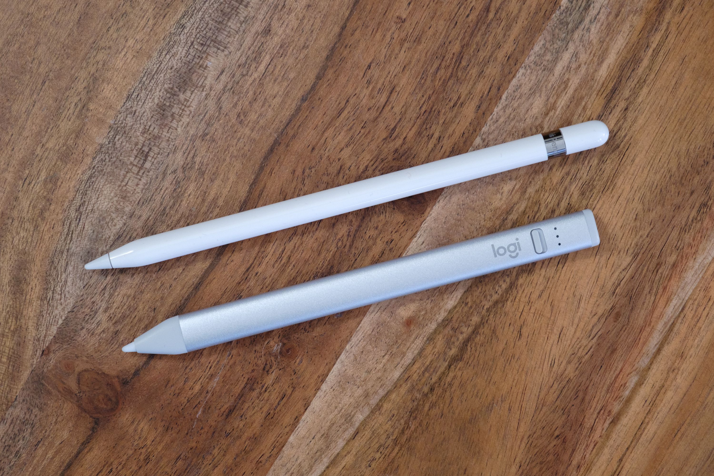 Logitech Crayon next to the 1st generation Apple Pencil on a wooden table, top view.