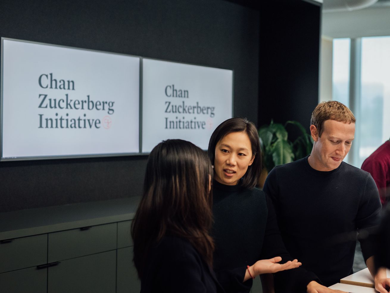 Priscilla Chan and Mark Zuckerberg speak with another person in front of a sign reading, “Chan Zuckerberg Initiative.”
