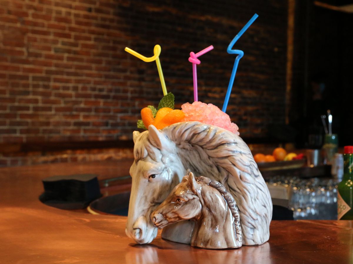Specialty bar glassware: a horse head with a pony head cozying up to it. A frozen cocktail and neon bendy straws are coming up out of the top. The glassware sits on a curved wooden bar with a brick wall in the background.