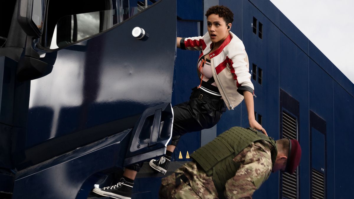 Ramsey (Nathalie Emmanuel) in Fast X hangs out of the driver’s-side door of a moving security truck, one hand on a uniformed soldier she’s just yanked out of the truck and is tossing to the ground.