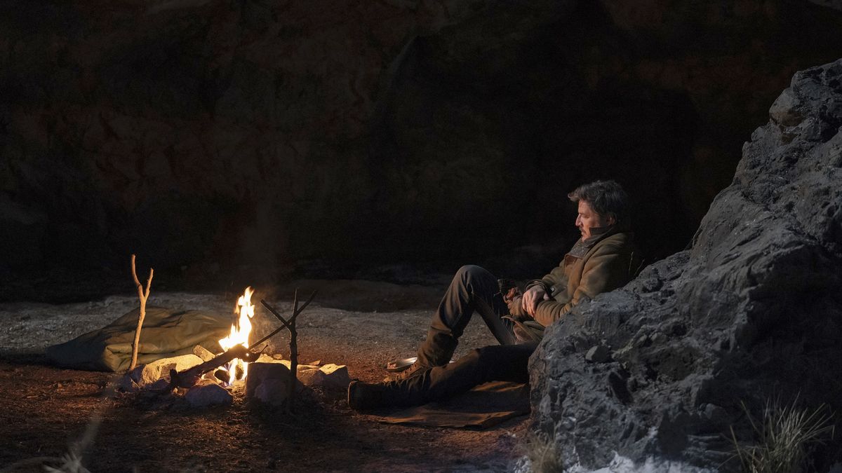 Joel (Pedro Pascal) sitting and leaning against a rock wall and looking at a fire