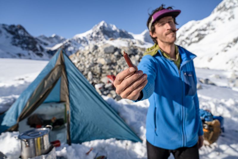 A person wearing Patagonia gear stands in front of a tent and holds out a piece of jerky towards the camera. There is snow on the ground and the mountains in the background are covered with snow.