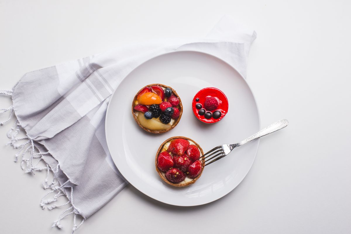 Three perfectly plated French custard tarts at Saint-Germain Bakery in Atlanta topped with blueberries, strawberries and raspberries on a crisp white plate atop a gray and white fringed napkin. A metal fork lays upside down, gently placed against the strawberry tart.