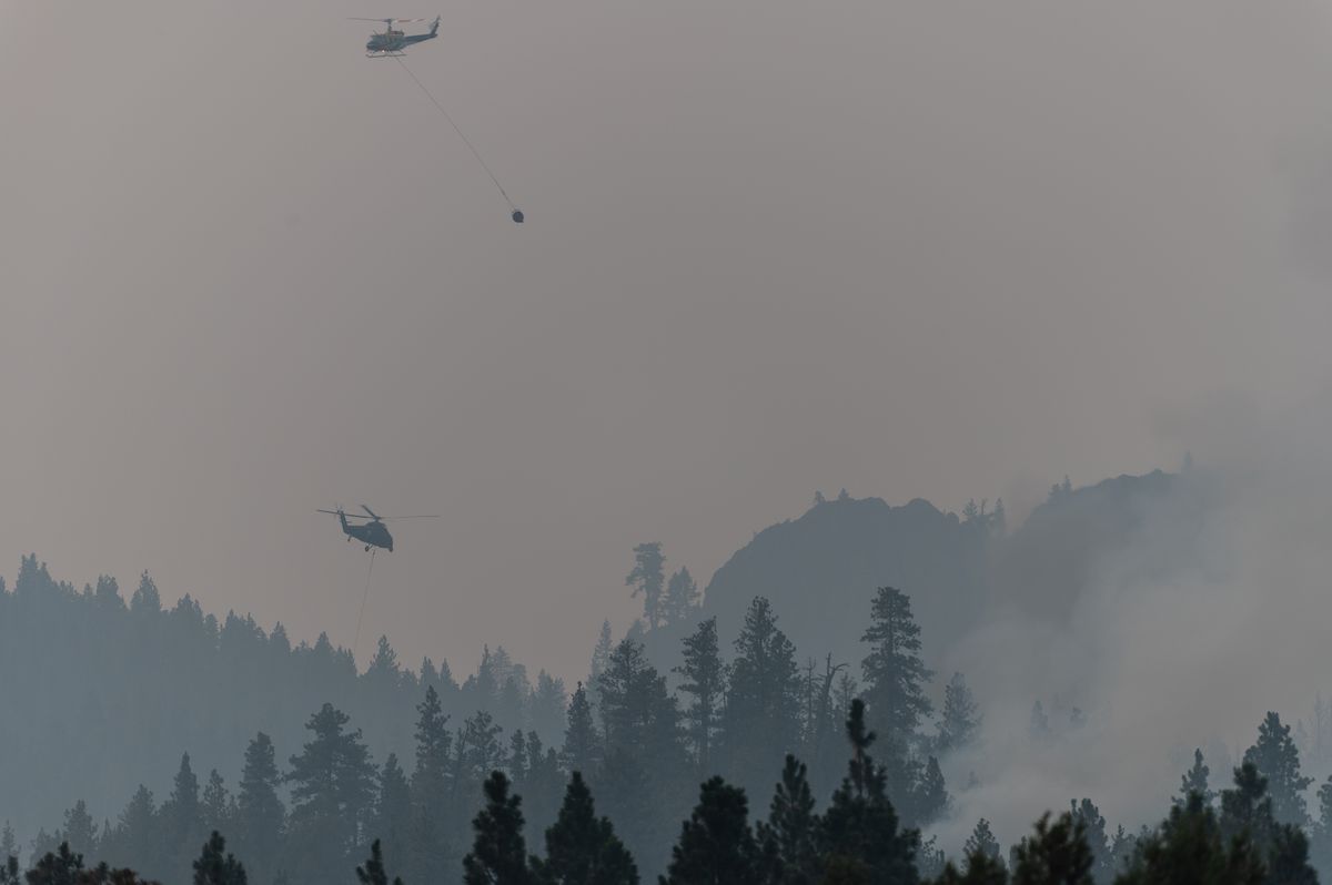 A helicopter flies above a tree line. The air is gray and hazy with smoke. 