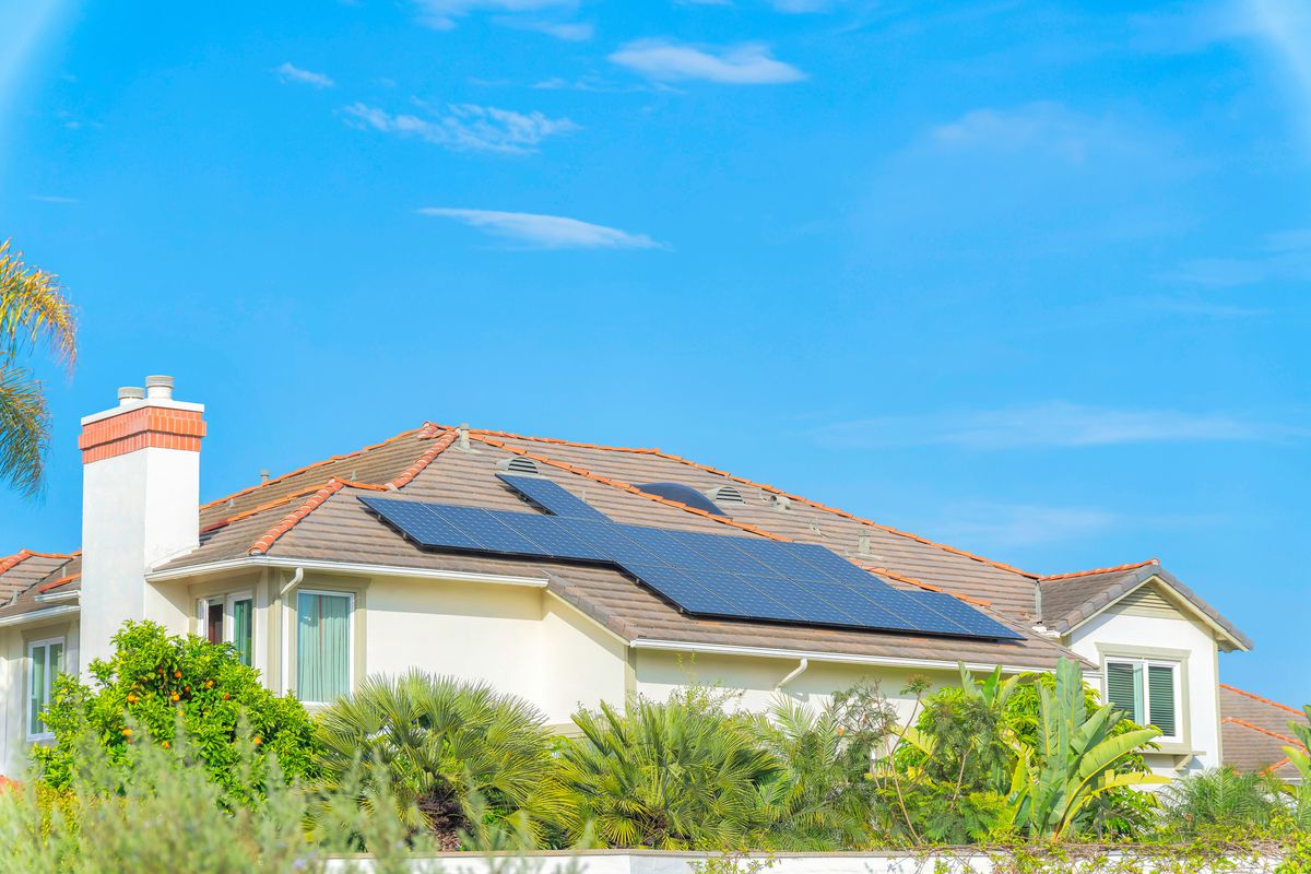 Solar panels on top of the roof of a house at Carlsbad, San Diego, California. Fenced house with trees at the front against the sky background.