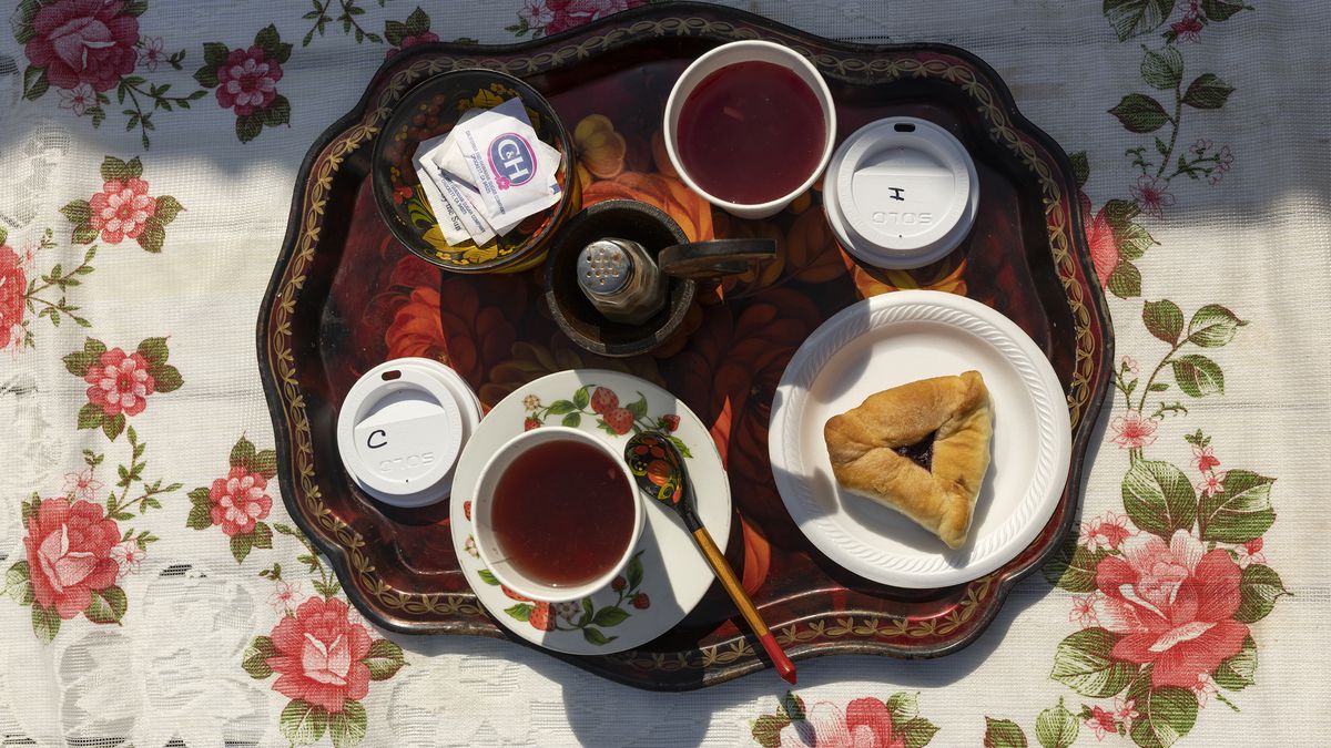 An overhead picture of a tray holding a cup of tea with a triangular pastry, sugar, on a floral tablecloth. 