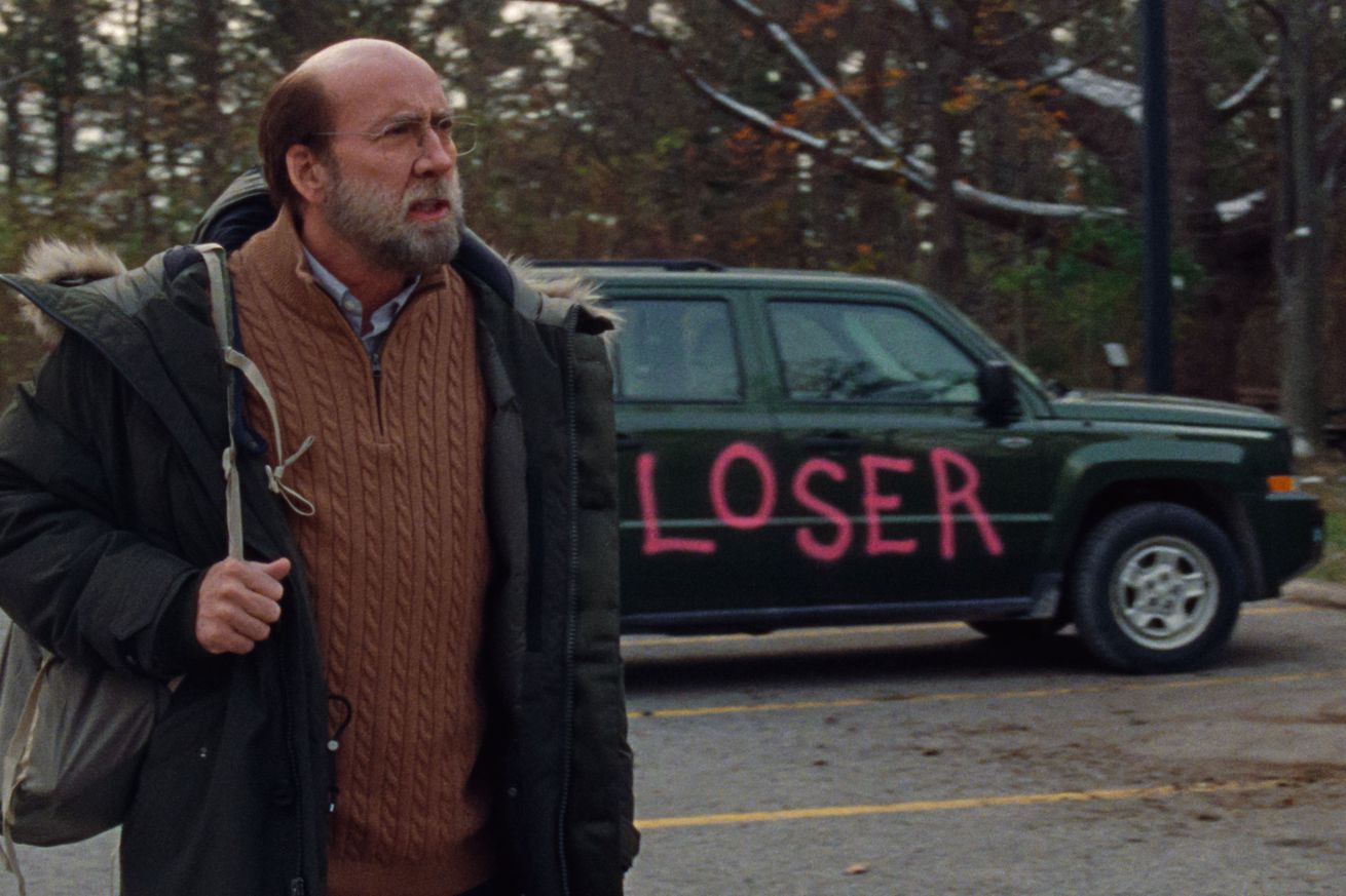 A bald man wearing a sweater and puffer coat standing next to a black sedan that has “loser” spray-painted on the side.