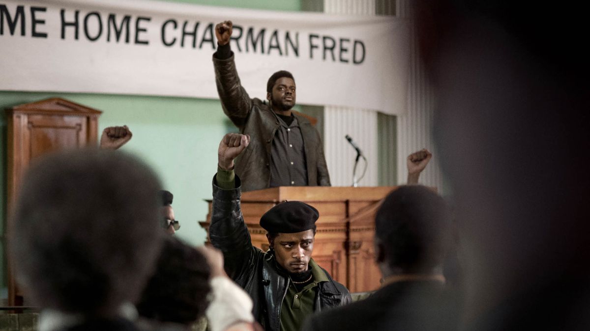 Fred Hampton stands at a podium in front of a banner reading “Welcome Back Chairman Fred,” with William O’Neal in front of him. Both raise their fists.