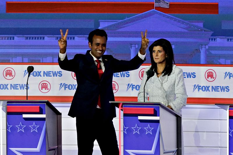 Vivek Ramaswamy and Nikki Haley on the debate stage. Ramaswamy holds up peace signs on each hand while Haley is blank-faced behind her podium.
