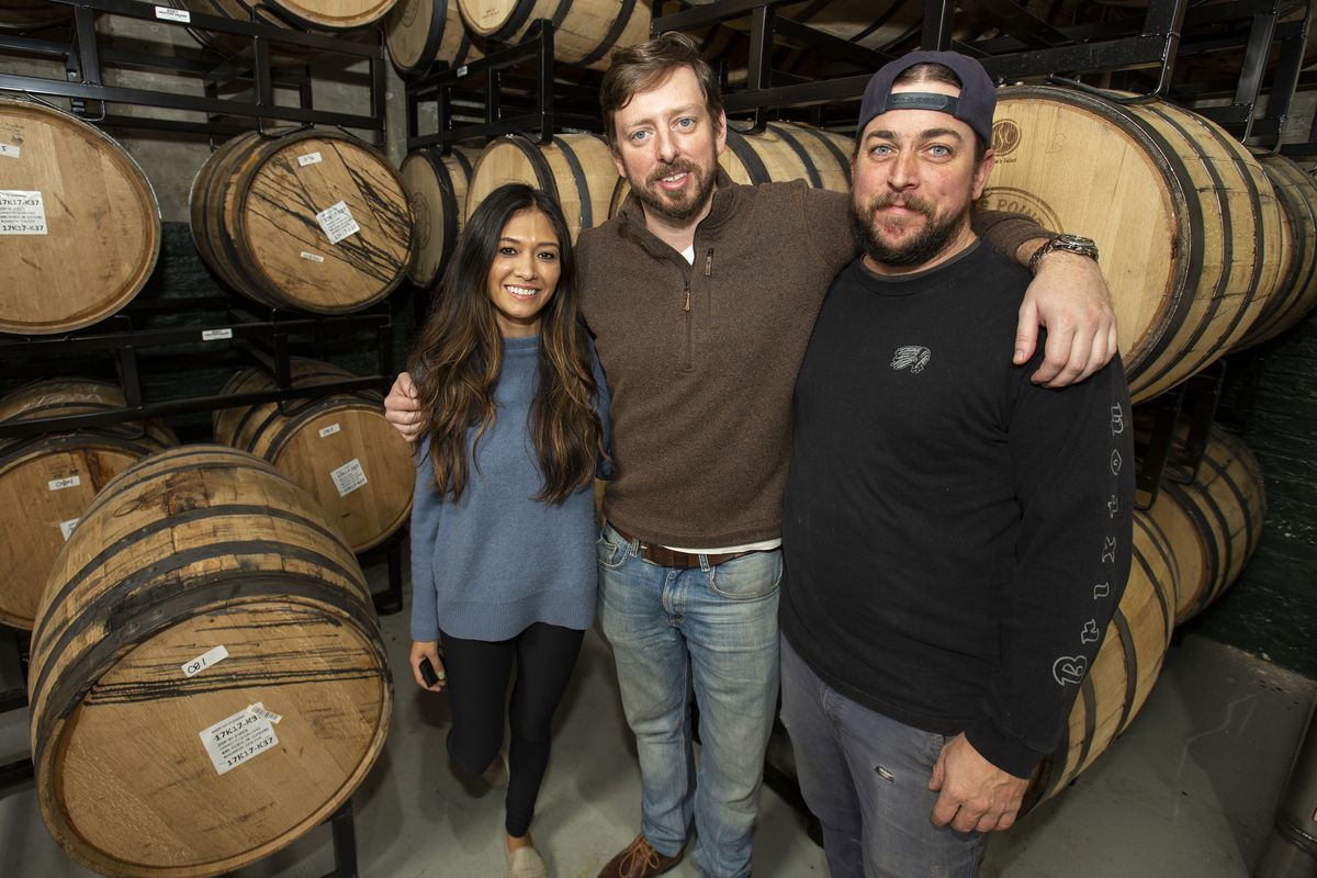 A woman and two men pose in front of large whiskey barrels.