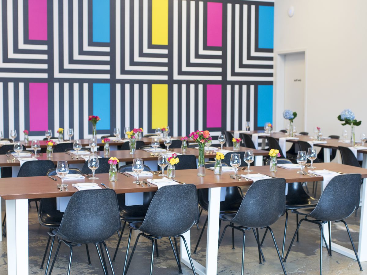 interior of aslin brewery with a colorful, abstract, geometric back-drop on the wall and black chairs situated around white tables.