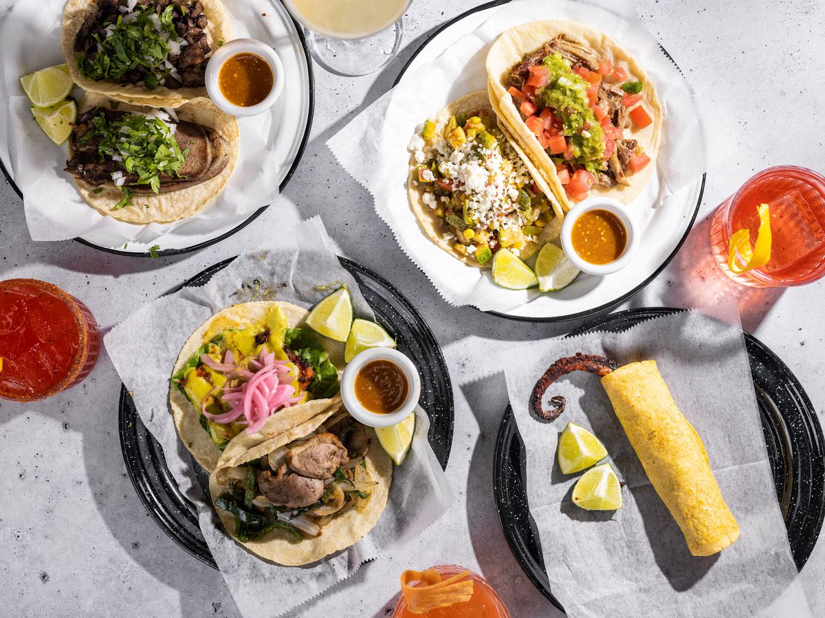Plates of tacos sit amid drinks on a white table.