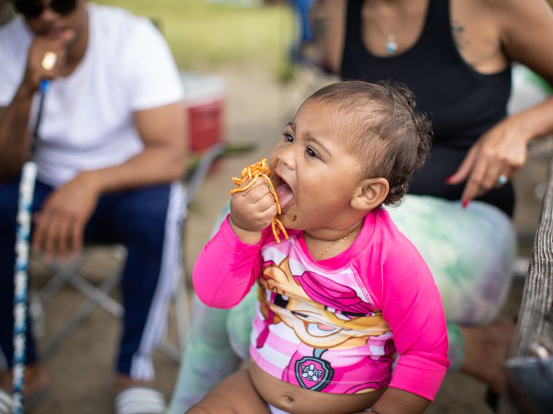 a baby in a pink swimsuit holds a fist of spaghetti to her mouth. 