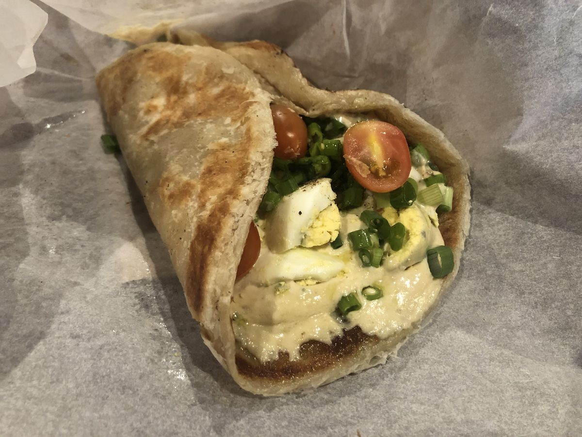 A fried pita flatbread wrap with hummus, tomatoes, and green onions inside, held in a paper wrapping