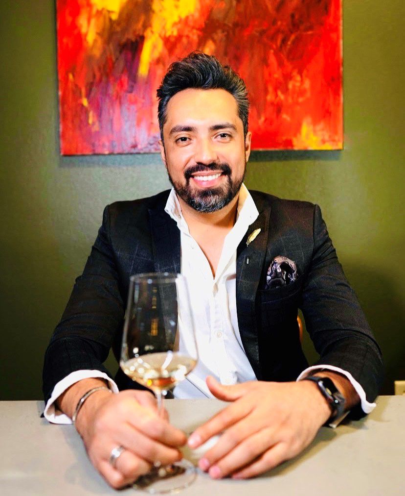 A man in a black jacket and white shirt holds a glass of white wine.