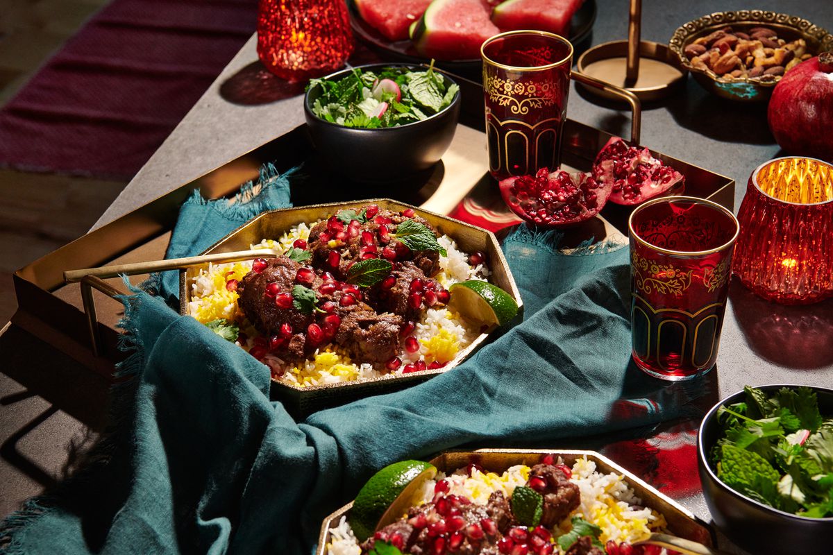 A big bowl of fesenjan on rice sits on a table surrounded by a lavish spread of sliced watermelon, glasses of wine, bowls of fresh herbs, and fresh pomegranates.
