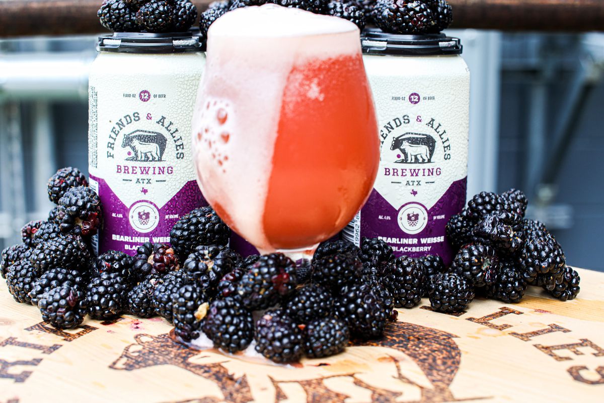 A glass overflowing with pink beer liquid with a pile of blackberries and beer cans.