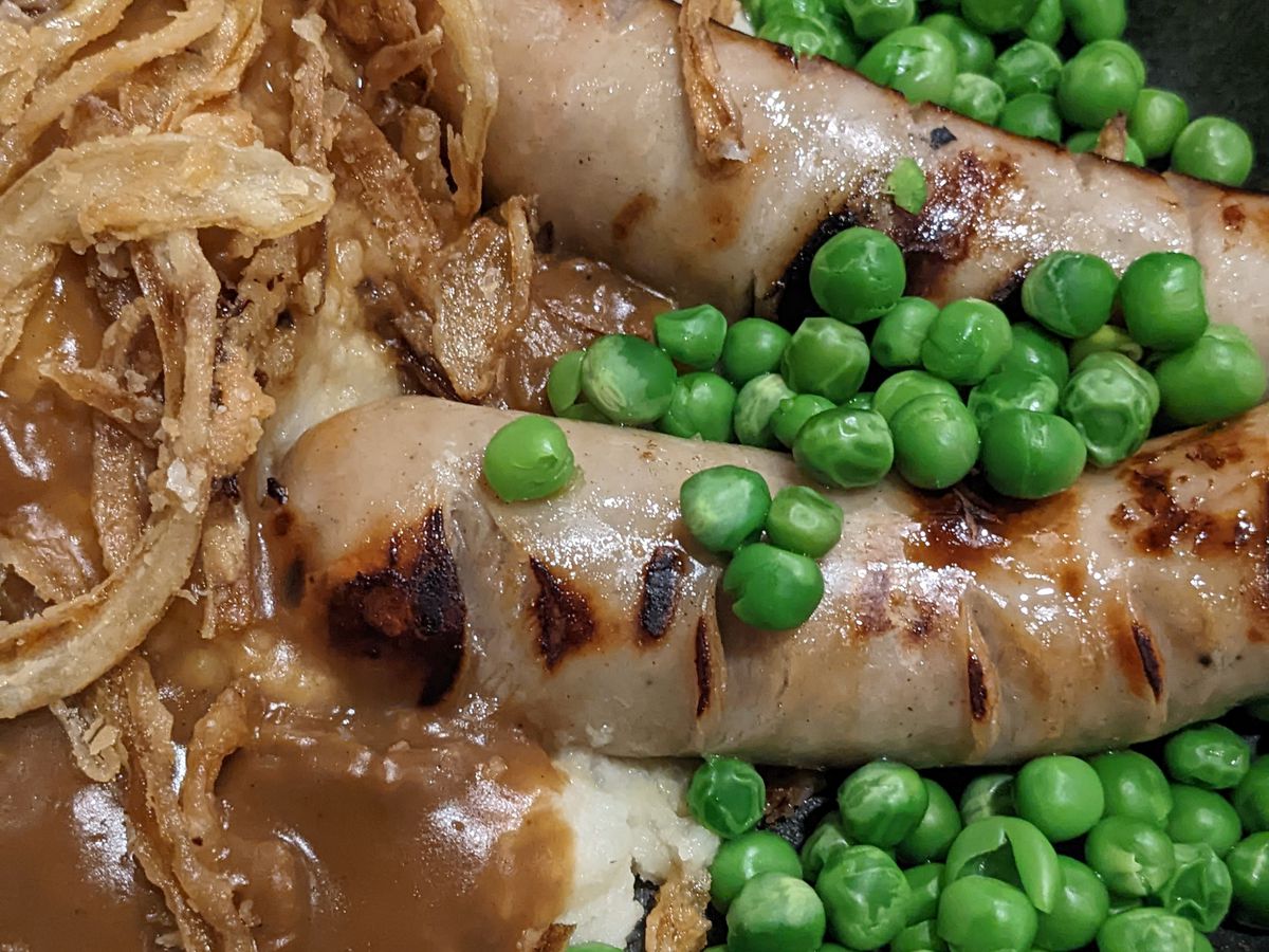 Two sausages atop mashed potatoes flanked by peas and fried onions.