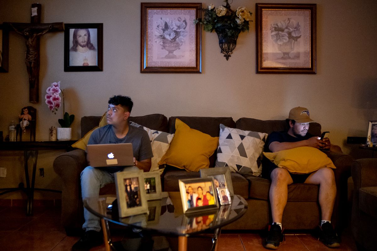 Two brothers sit on opposite sides of the couch, one doing schoolwork on a Mac laptop while the other looks down at his phone. The walls and tables of the living room are adorned with pictures, flowers and a large crucifix.