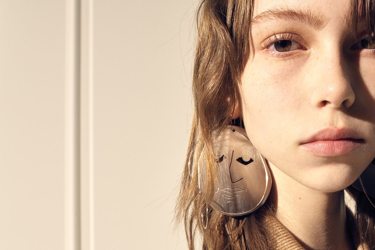 A model wears a pair of circular translucent earrings with a face sketched on them