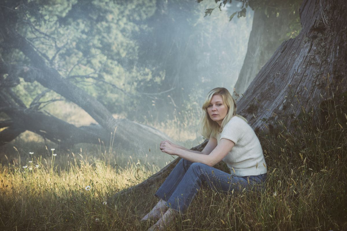 Dunst sits at the base of a tree, wearing blue jeans and a white knit t-shirt.