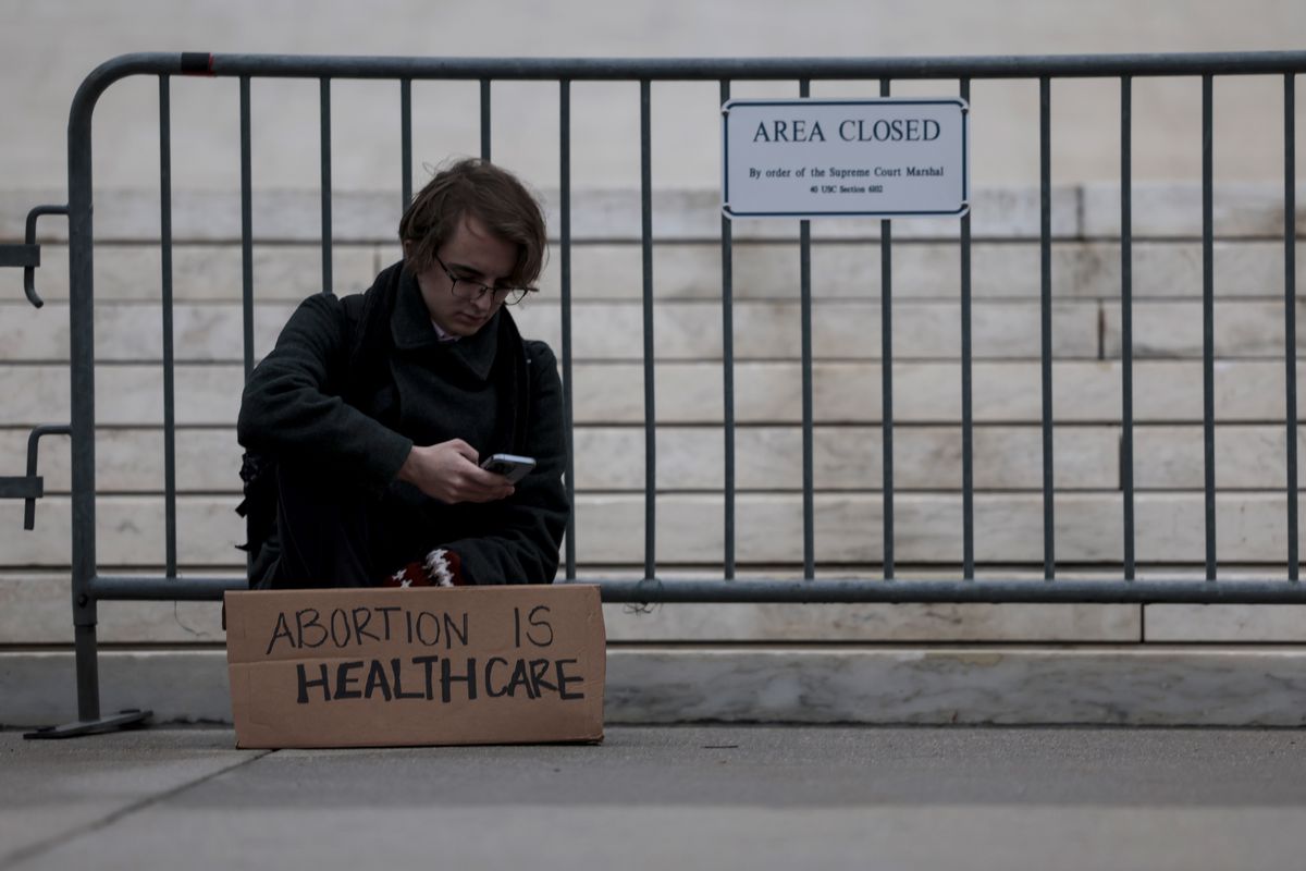 A person with an “Abortion is health care” sign sits looking at their phone in front of a barricade outside the Supreme Court.
