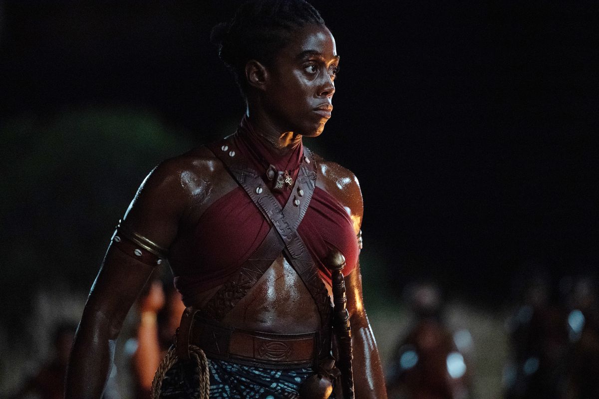Lashana Lynch standing in her red top leather straps and sword hilt ready for battle