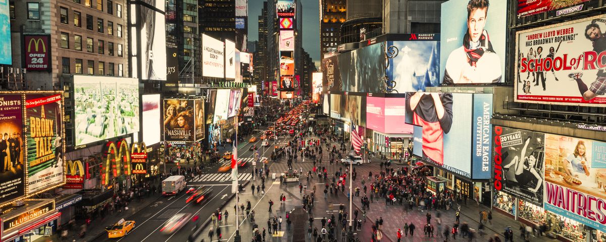 An aerial view of the Time Square pedestrian mall surrounded by many illuminated billboards and signs on buildings. There are people walking in the mall area.