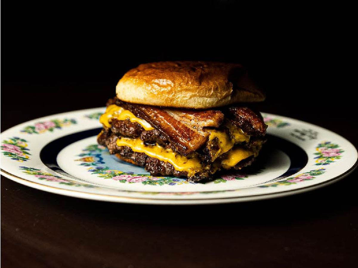 A double smash cheese burger topped with bacon on a round white plate with a floral pattern, against a black background. 