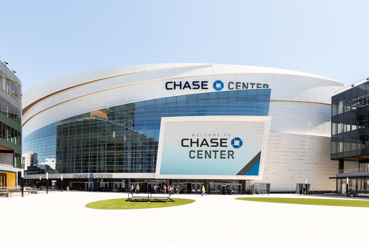 Large, white circular building with white perforated panels. A large series of windows cuts across the front of the building, with a large display reading “Chase Center” attached to the front.