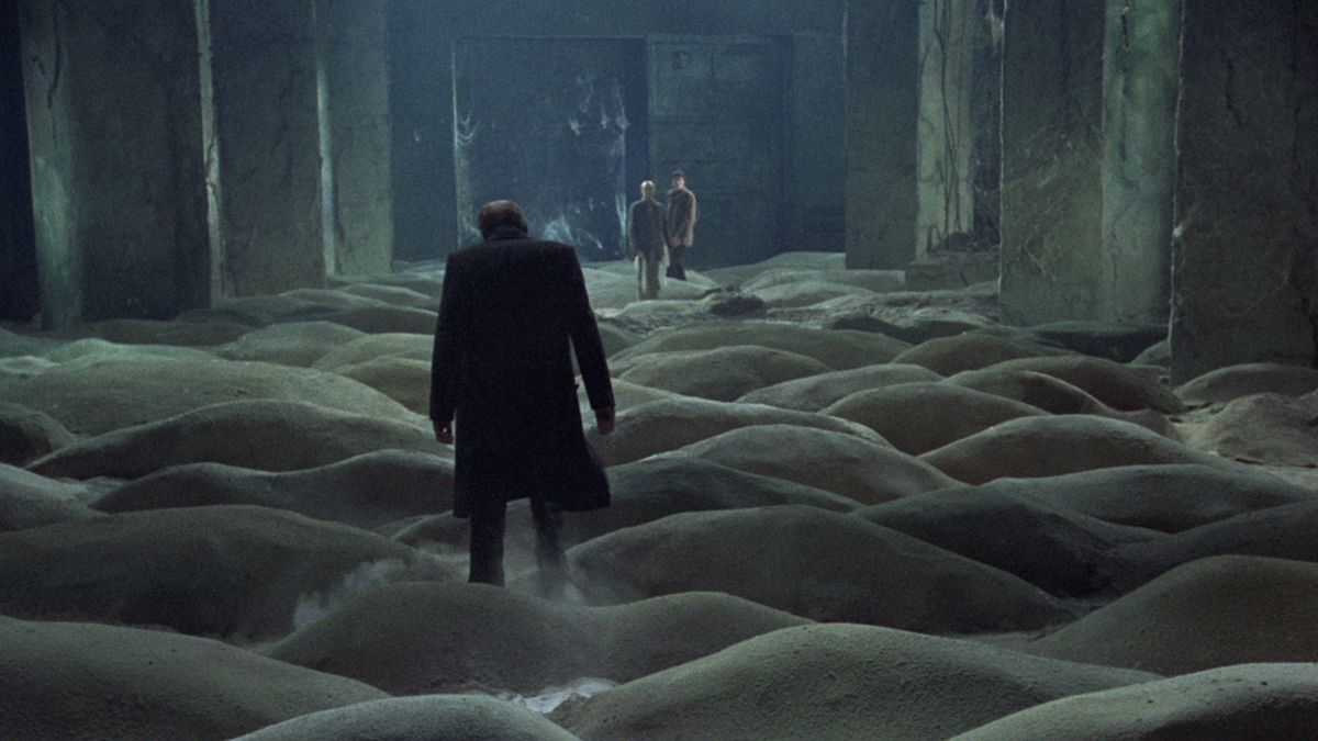A man in a black coats walks through a room covered in mounds of dirt with two other men at the far end standing beside one another.