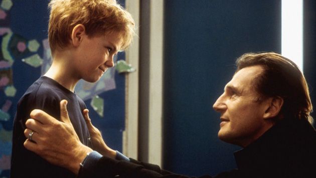 liam neeson kneels in front of a young boy, urging him to confess to his crush