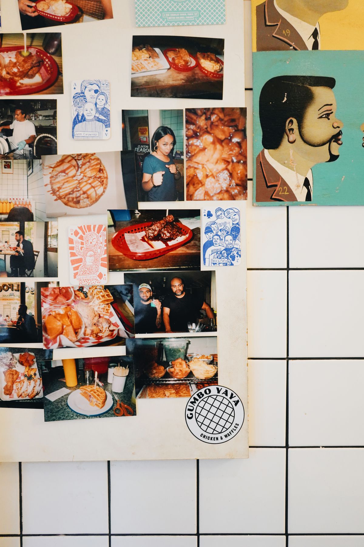 A close up of a colorful collection of photographs and drawings of people and food, all tacked up on a white tile wall inside Gumbo Yaya.