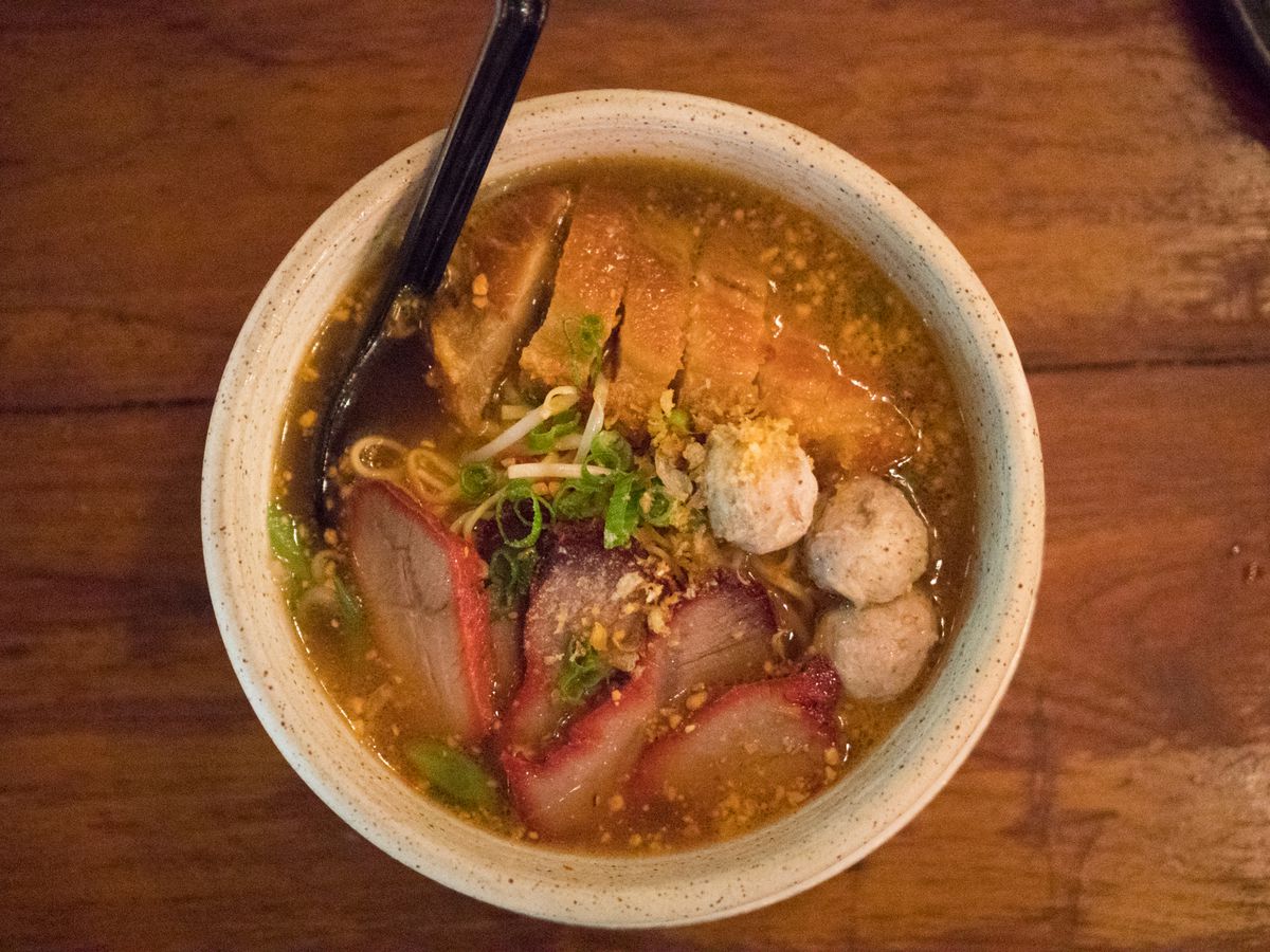 A white bowl of Ba Mhee “Pitsanulok,” a noodle soup with meatballs, pork, and chunks of chicken. The bowl is served on a wooden table with a black spoon, with dots of crushed peanuts in the broth.