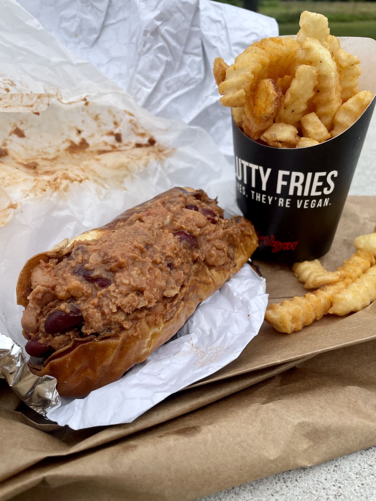 Hot dog covered with chili and served with french fries.  They both sit on a foil wrapper with a brown paper bag on the right side. 