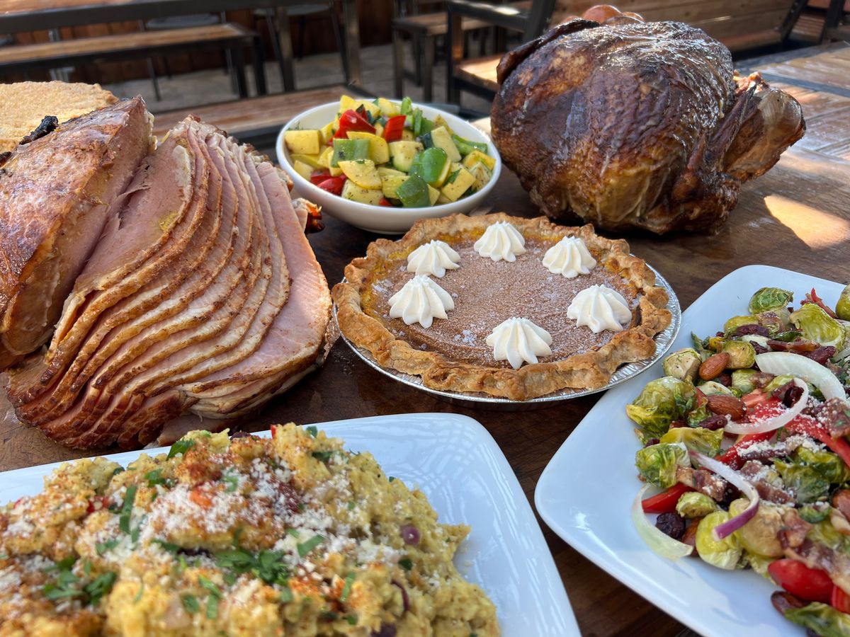 An assortment of barbecue dishes and Thanksgiving dishes are laid out on a wooden picnic table outdoors.